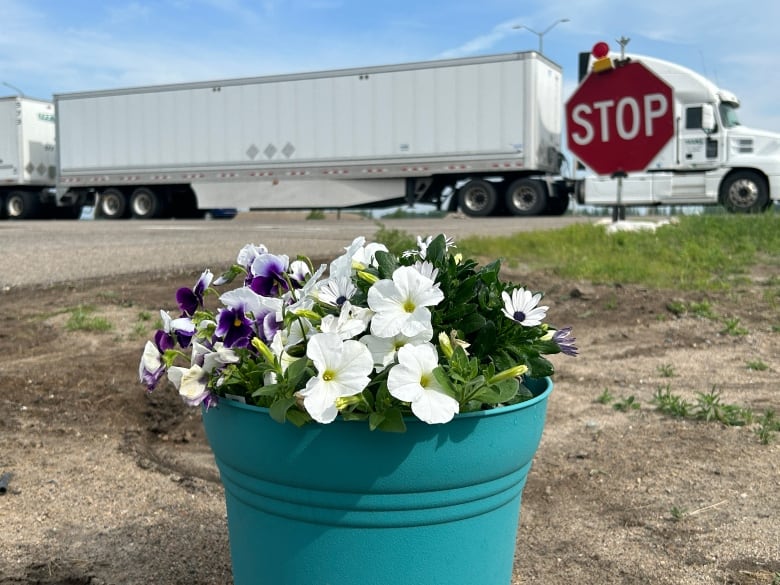 A pot of flowers is in the close-up foreground with a stop sign in the background and a semi-trailer passing by on the highway.