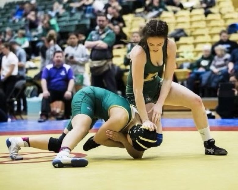 Two women in green leotards participate in a wrestling match inside a school gymnasium.