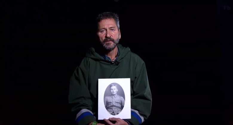 A man standing in front of an all-black background. He's holding a black-and-white portrait of a soldier, and he's wearing a traditional Inuit cossack.