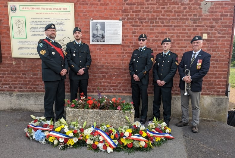 Five men in military garb standing in front of a plaque on a brick wall.