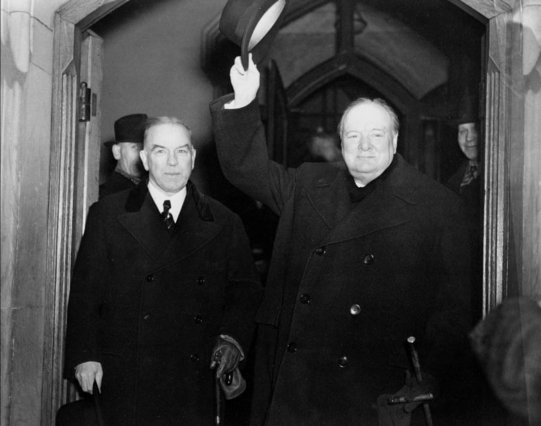 Sir Winston Churchill raises his arm in the air holding a hat while Prime Minister William Lyon Mackenzie King walks beside him, leaving the House of Commons.