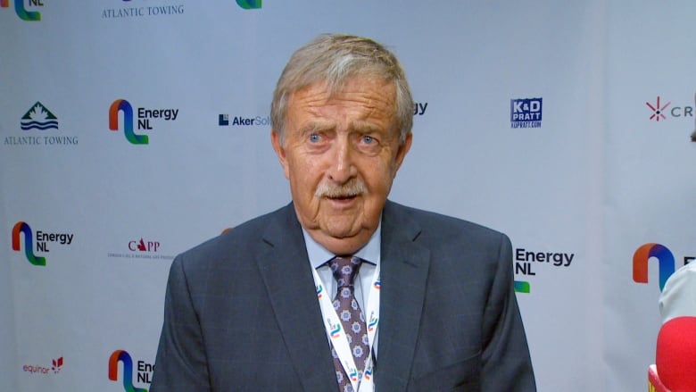 An older man with grey-brown hair and a grey moustache. He's wearing a blue suit and standing against a background with logos for Energy NL and CAPP on it.