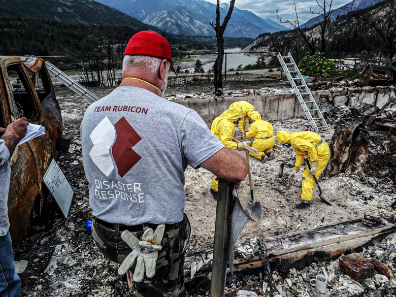 A man in a red ball cap and a grey shirt that says Team Rubicon Disaster Relief on its back watches three people in yellow hazmat suits work amid the rubble.