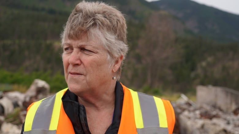 A woman with short brown and white hair, in a high-vis vest, stands among rubble.
