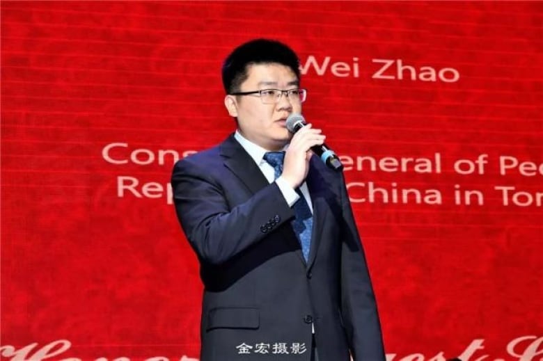 A man in a suit, standing against a red backdrop, speaks into a microphone.