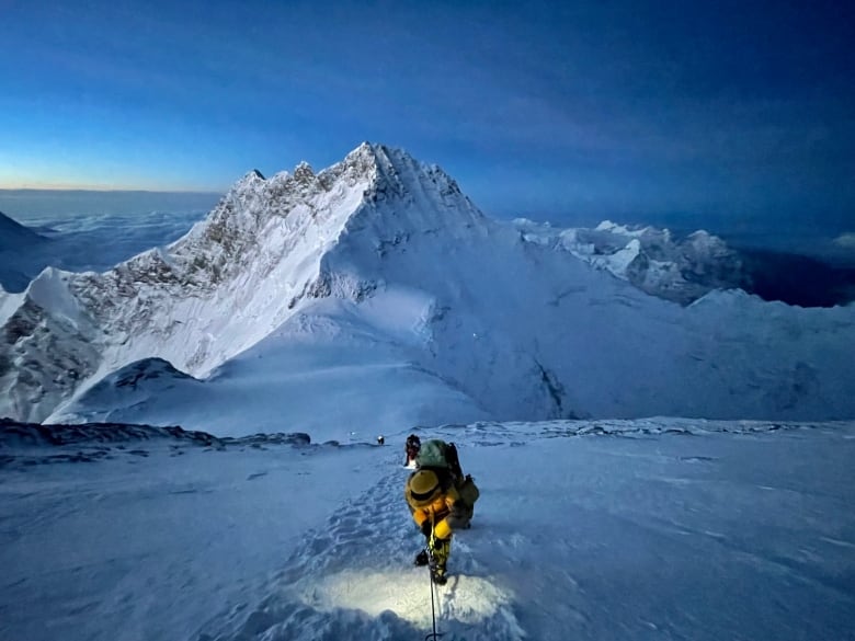 A person can be seen climbing up a snowy mountain with another mountain in the background.