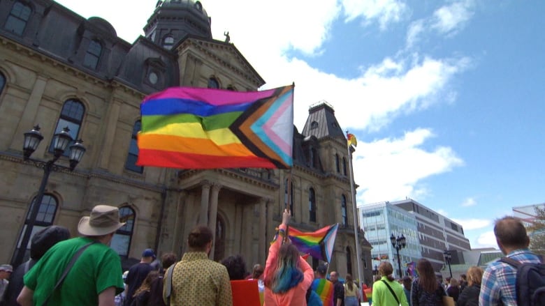 People holding up pride flag in foreground, legistlative assembly in background.