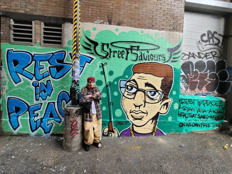 A man stands in front of a mural on an alley wall of a man with a halo and "Rest in Peace" written beside it.