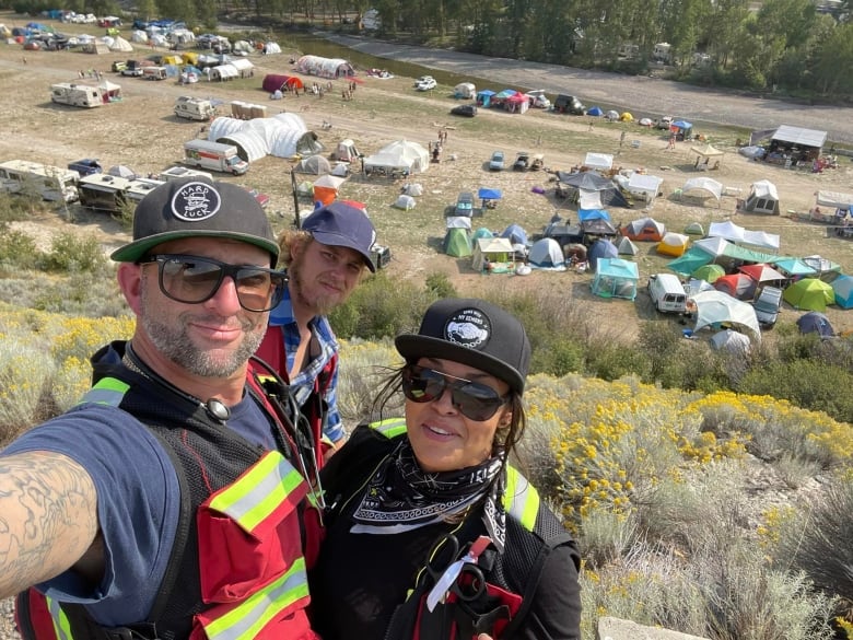A selfie of a man and two others in medic vests with tents on a field below.