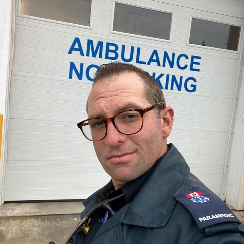 A man in glasses and a paramedic uniform smiles, with an ambulance parking sign behind him.