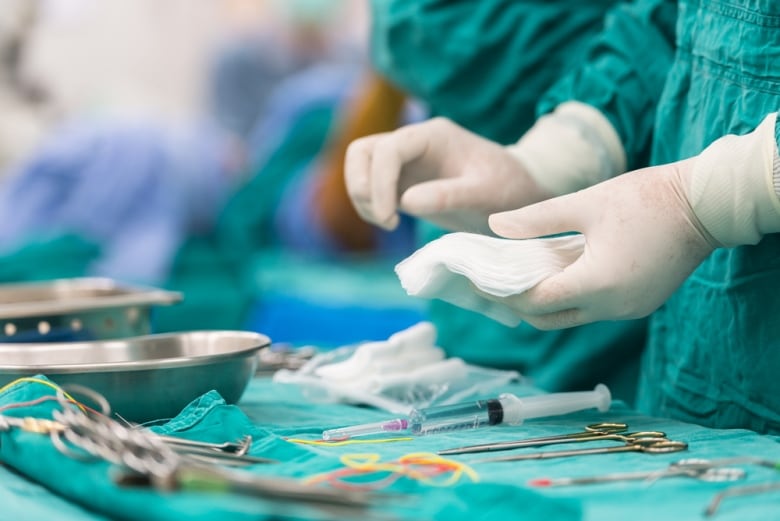 A closeup photo shows the gloved hands of a health-care worker in green surgical scrubs, holding a piece of gauze above an array of surgical tools.