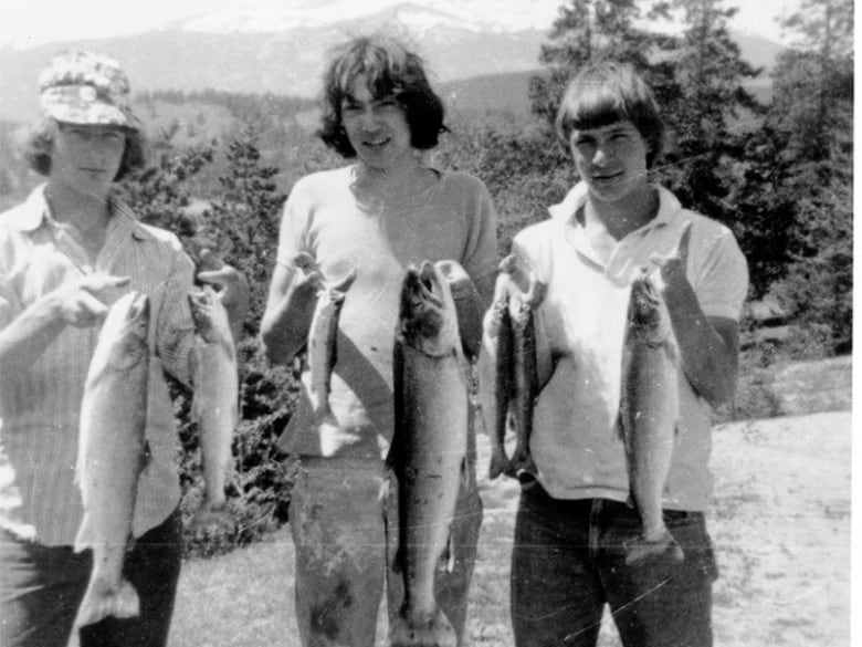 Three men hold fish and stand together with mountains and trees in the background.
