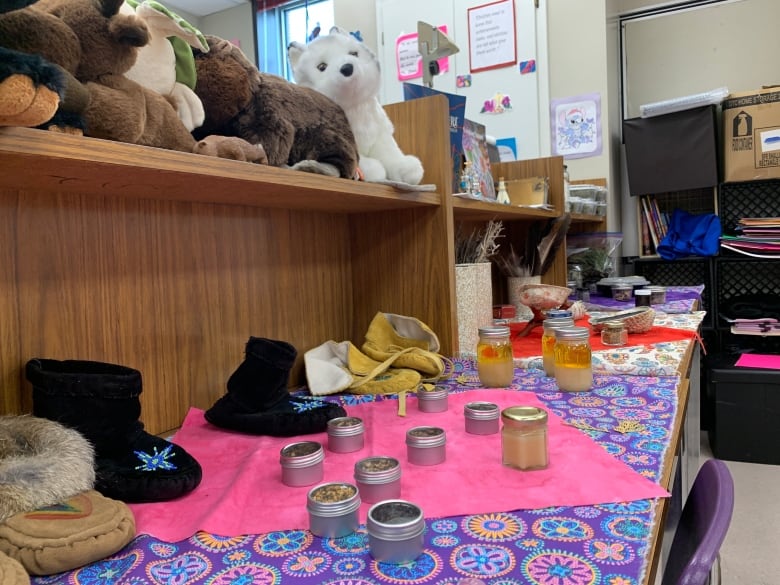 Moccasins and little vials are seen on top of brightly coloured fabric laid out across a table. Above, stuffed animals are perched on a shelf.