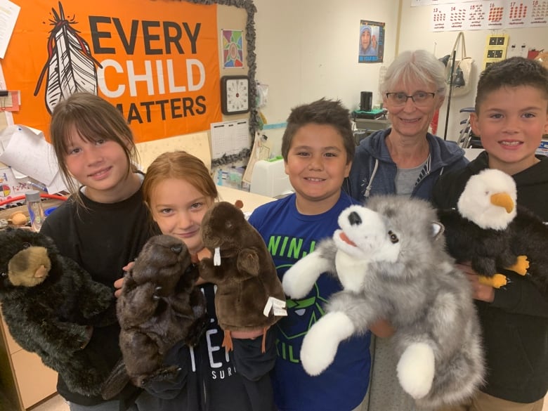 Four children hold up stuffed animals in a classroom with woman in glasses and white hair. In the background is an orange flag that says, "Every Child Matters."