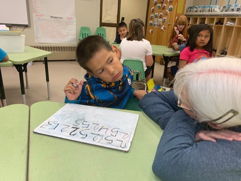 A woman with white hair kneels next to a little boy who is writing numbers on a little white board.