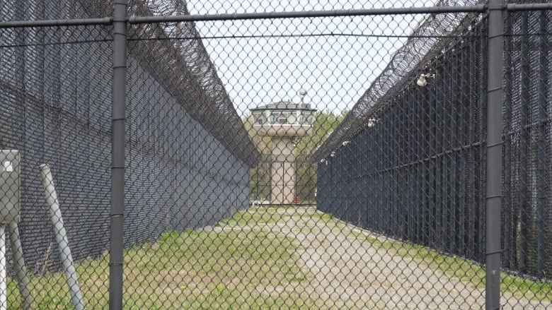 A guard tower can be seen through a black, chain-link fence. There are two lines of fencing wrapped in barbed wire running parallel up to the tower.