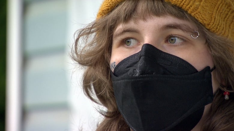 A closeup shows a person wearing a black medical mask. They have hazel eyes, a silver eyebrow ring, curly brown hair and a mustard yellow toque.