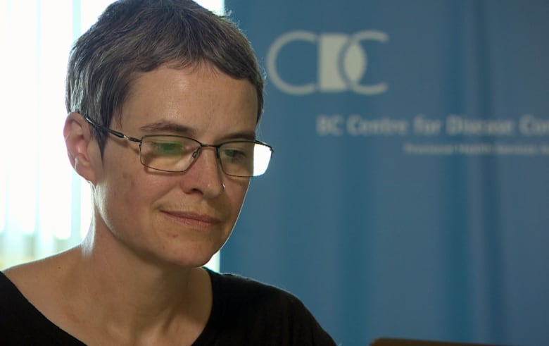 A white woman with short cropped grey hair and wire-rimmed glasses looks down at a laptop in front of a blue banner reading B.C. Centre for Disease Control.