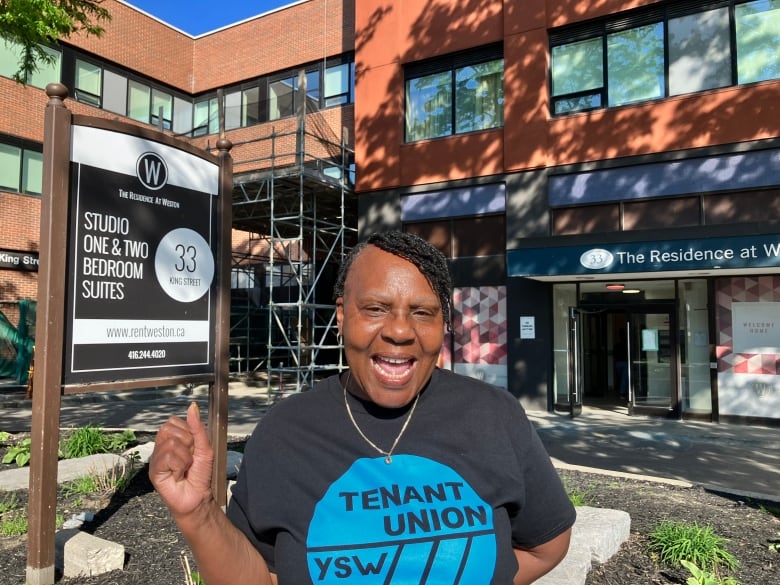 A woman in a short that says Tenant Union stands near a sign advertising apartments in her building.