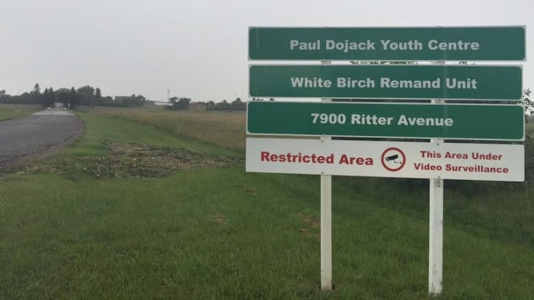 A large green and white sign is shown on a patch of grass, next to a road. It reads: Paul Dojak Youth Centre. White Birch Remand Unit. 7900 Ritter Avenue. Restricted Area: This area under video surveillance.