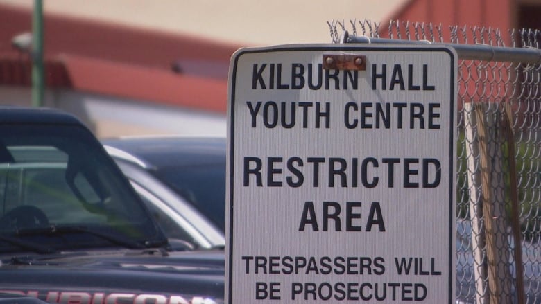 A black-and-white metal sign is affixed to a chain-link fence, with vehicles parked behind. It reads: Kilburn Hall Youth Centre. Restricted area. Trespassers will be prosecuted.