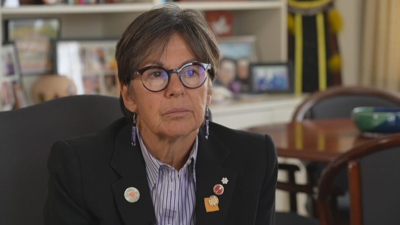 A woman wearing black-rimmed glasses, a black blazer and a striped shirt sits in an office chair, with framed photos and a table shown behind her.