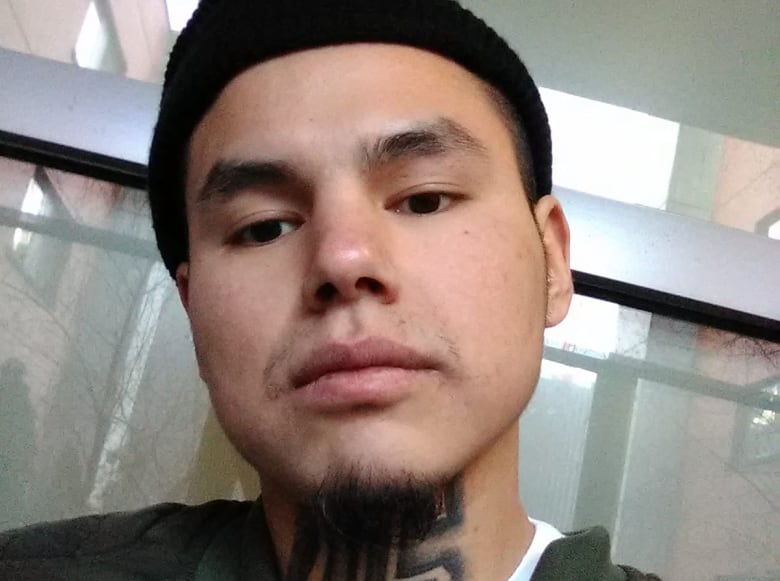 A young man wearing a black beanie, white T-shirt and black hooded sweatshirt poses for a selfie in front of a window. He has a neck tattoo of a 'G' with two vertical lines running through it.