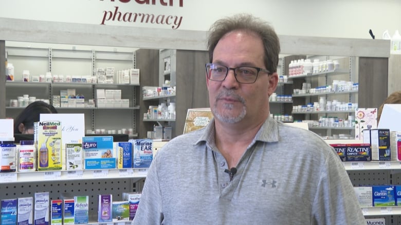A man in a grey shirt stands in front of a pharmacy counter. 