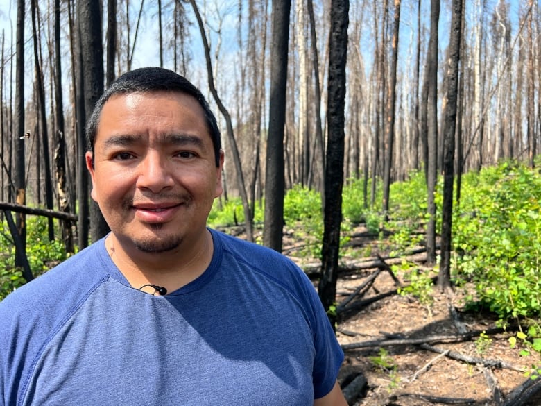 A man looks at the camera, smiling. He wears a plain, navy T-shirt. Behind him, you can see a forest with charred trees and some green regrowth on the ground.