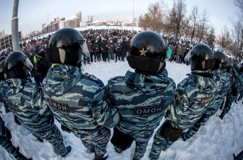 Russian police in black helmets watch protesters in the snow.