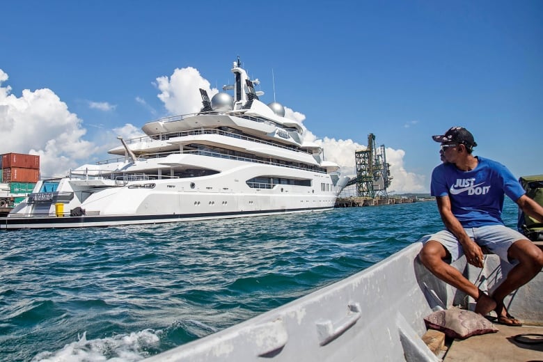A man in a blue shirt and baseball cap looks out at a large yacht in the ocean. 
