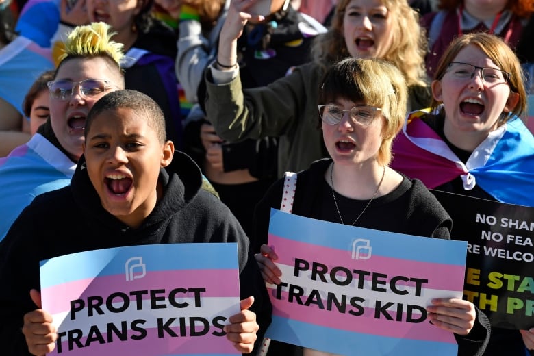Teenaged kids hold pink and blue striped signs that say "Protect Trans Kids" 