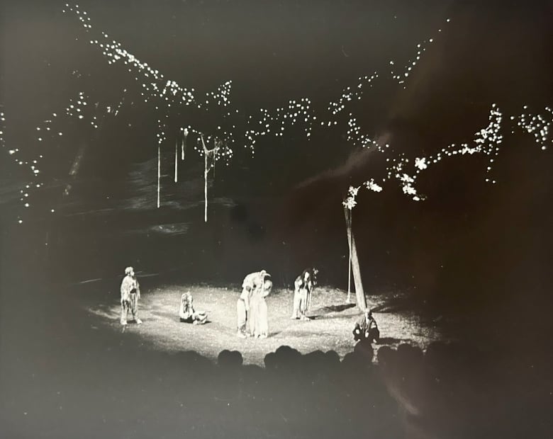 Black and white photo of actors performing at the High Park amphitheatre with lights strung from trees overhead.
