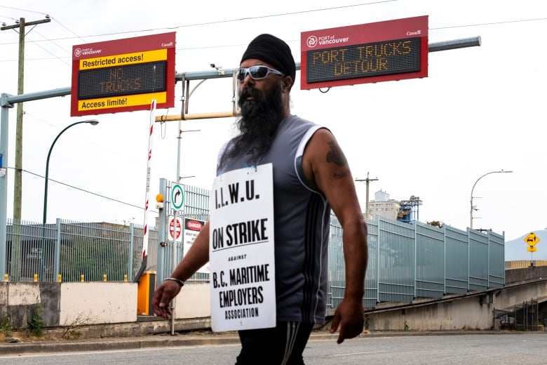 A South Asian man with a flowing beard walks on a street wearing a sandwich board that reads 'I.L.W.U. On Strike against B.C. Maritime Employers Association'.