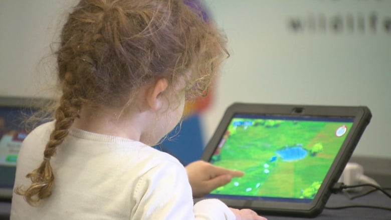 A little girl with a braid stands in front of a tablet playing a computer game.