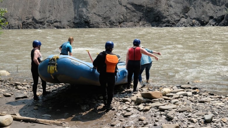 People in lifejackets and helmets place a blue raft into a river. 