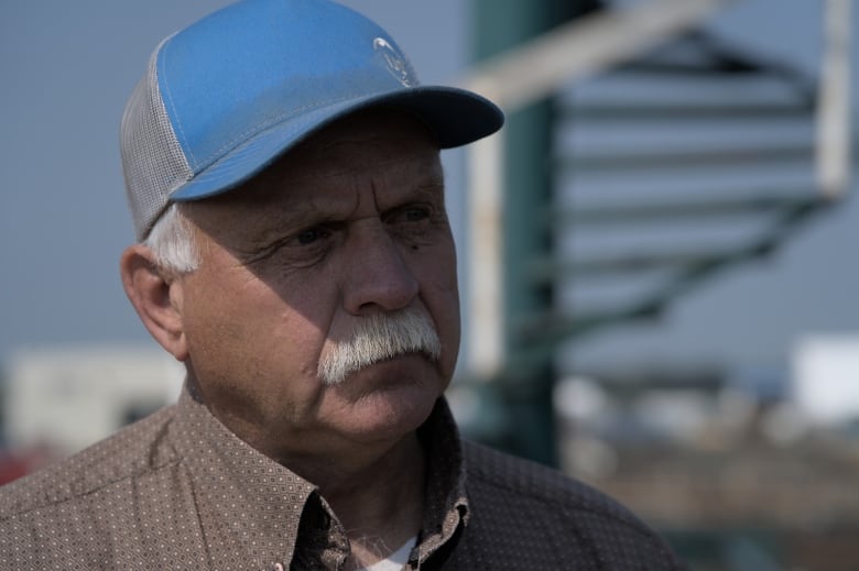 Older man with grey mustache and ball cap standing above pens of cattle. 