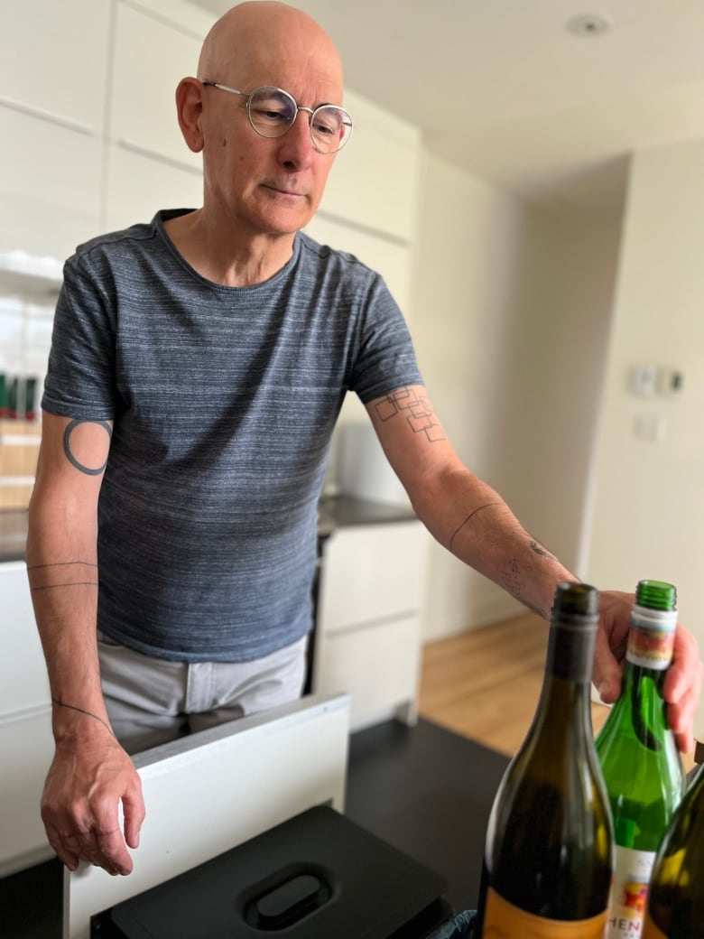 A bald white man with minimalist tattoos recycling wine bottles in his kitchen.