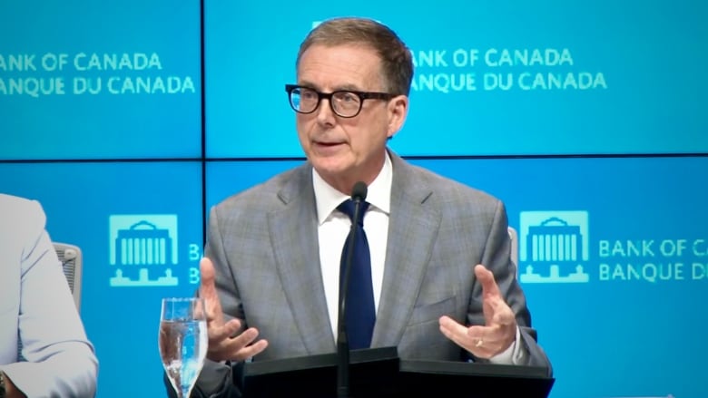 Bank of Canada Governor Tiff Macklem is pictured in a screen grab from an online video stream of the press conference. He's holding both hands up in front of him, as though grasping. There's a water glass on the table in front of him and a blue background with multiple "bank of canada" logos and titles.
