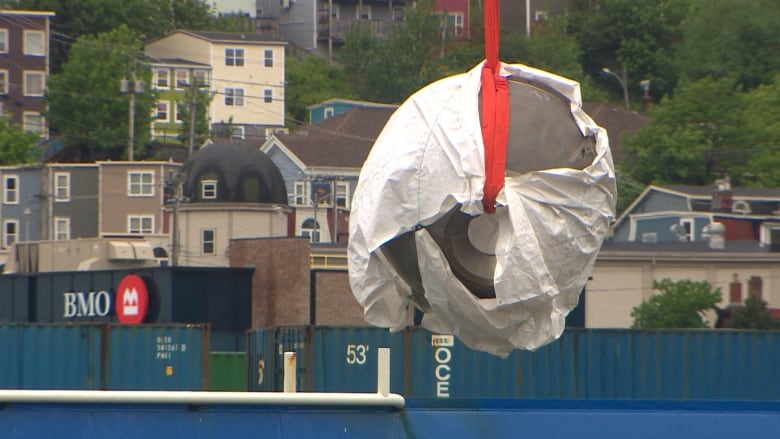 A large piece of semi circular titanium with a window in the middle. It's being lowered to the ground by a crane, and colourful houses of downtown St. John's are visible in the background.