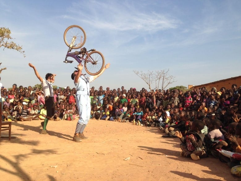 A man dressed in blue balances a bike on his face surrounded by children in a dirt field. 