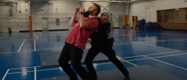 An police office has her arm wrapped around a volunteer to demonstrate the carotid control technique. The pair are in a gym and practicing on a mat.