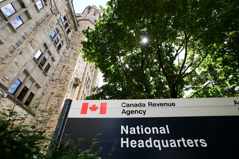 A sign reading "Canada Revenu Agency" under the shade of a tree stands next to an old brick building.