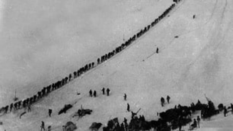Black and white photo of a line of people climbing a steep snowy mountain.