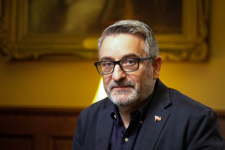 A man with grey facial hair and glasses looks into the camera. 