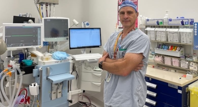 Dr. Craig Pearce is dressed in light blue scrubs and stands with his arms crossed in front of three computer monitors and anesthesiology equipment.