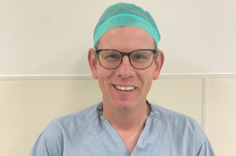 Dr. Trevor Rudge is wearing blue scrubs, a surgical cap and classes as he stands in front of a wall and smiles at the camera.