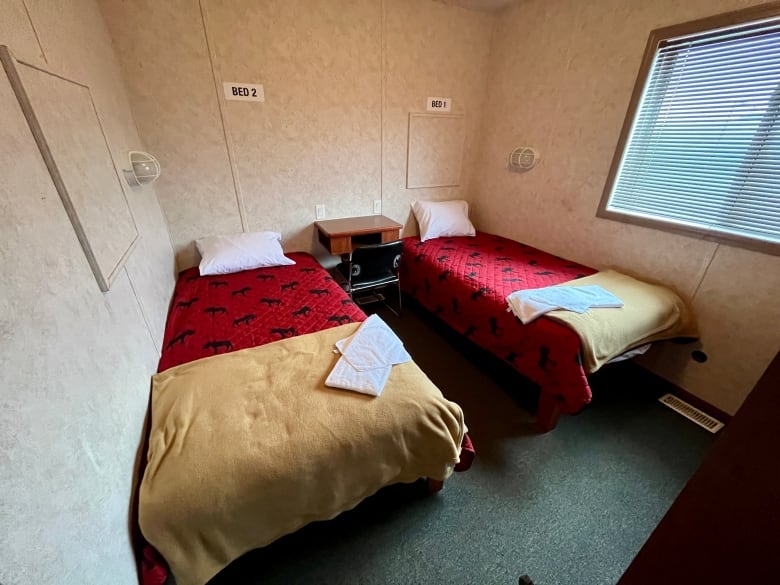 Two single beds side by side with red coverlets with a moose pattern, fleece blankets folded at the bottom, and towels folded on top. There is a window on one wall and a small table between the beds. 
