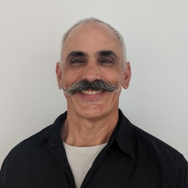 A man with a moustache smiles as he poses for a portrait in front of a white wall.