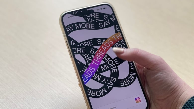 A hands with glittery nail polish hold a cellphone. On its screen is the looping logo of Threads, a social media platform. It reads: Threads: Say more.